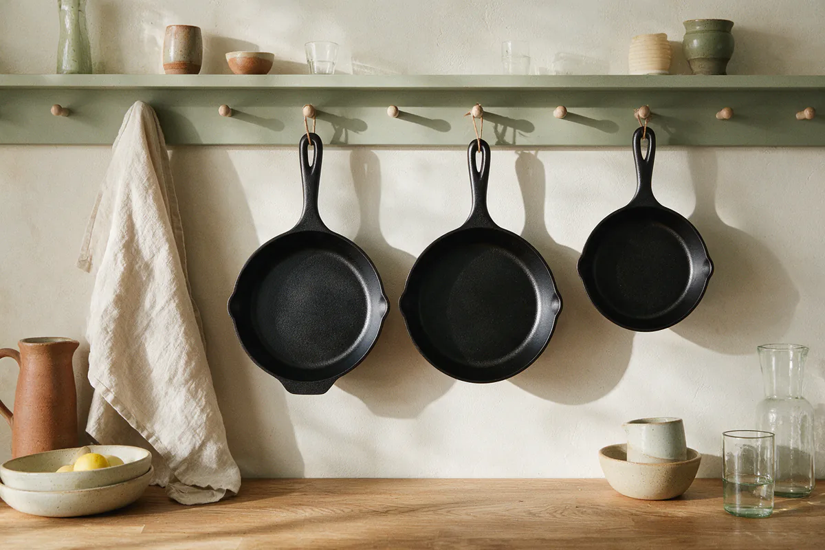 Three cast iron skillets of different sizes hanging on hooks above a wooden counter.